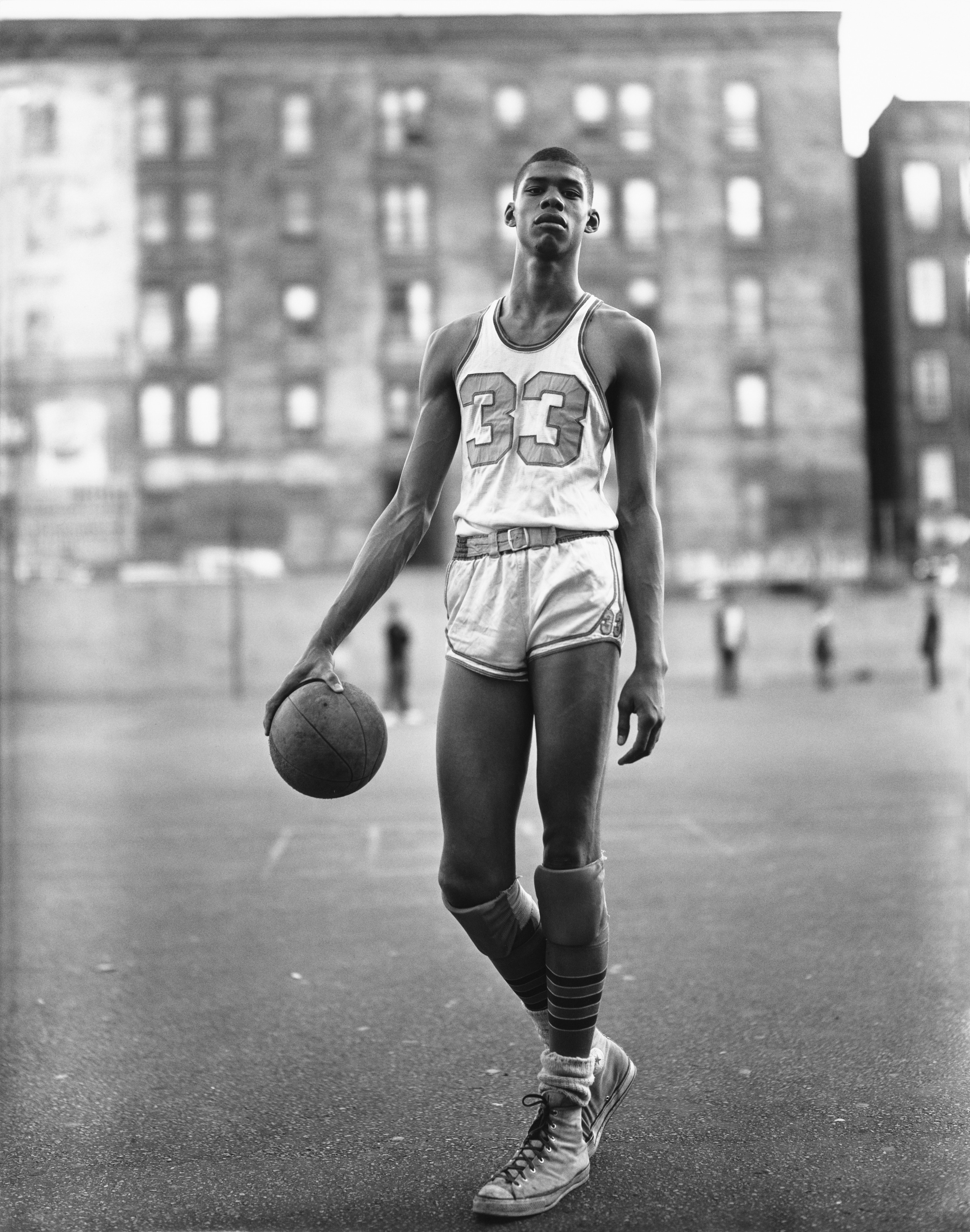 A young Lew Alcindor in his number 33 kit holds a basketball at his side on an outdoor New York court, tenement blocks visible behind him.