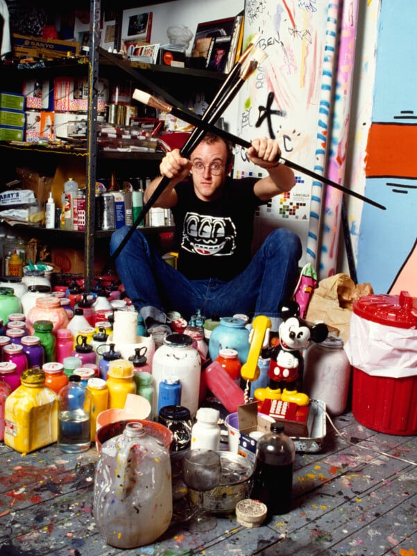 A man in glasses sits cross-legged on a paint-splattered floor, surrounded by colorful paint bottles and supplies, holding several paintbrushes above his head in a playful pose. Shelves and art fill the busy, creative studio.