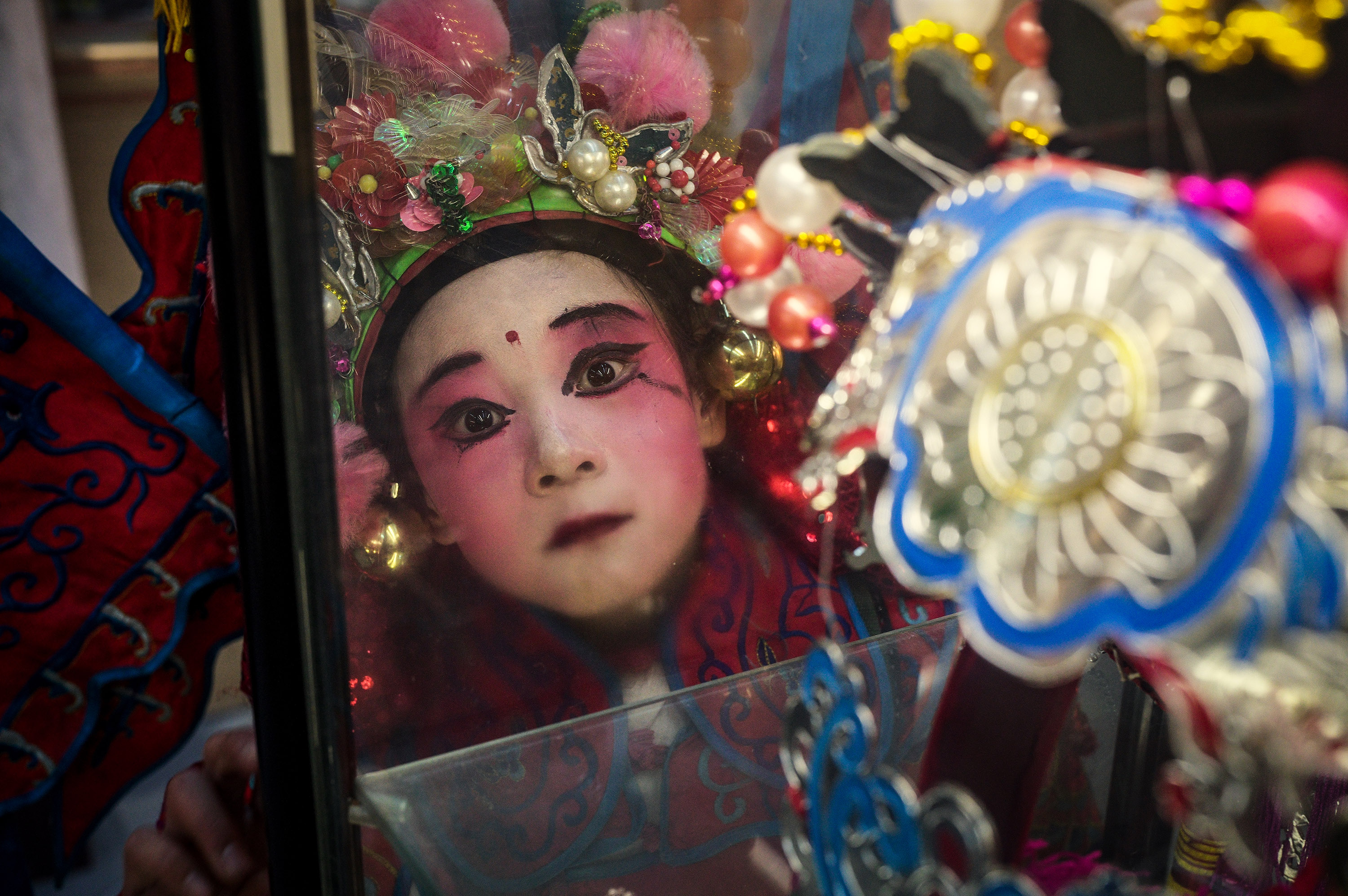 A child in red traditional costume with bright pink face makeup and decorated headdress gazes at her reflection in a mirror surrounded by elaborate props.