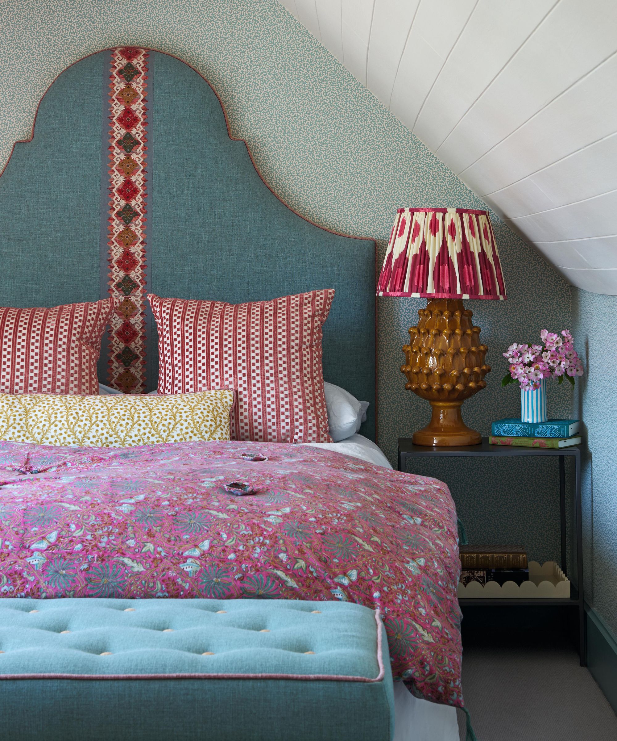a master bedroom with sloped paneled ceilings, ditsy print wallpaper, a curved headboard and pops of blue and pink