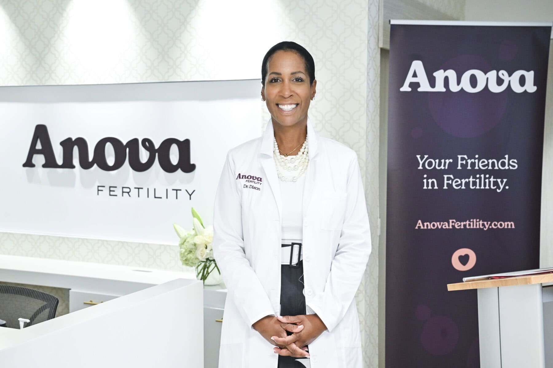 A smiling female fertility specialist in a white lab coat standing in front of Anova Fertility branding at a Toronto clinic, promoting fertility treatments and reproductive health services.