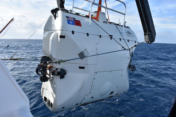 A white TRITON submersible is suspended above the ocean, attached to a crane on a ship. The sub has an American flag sticker and is being lowered or lifted from the water under a partly cloudy sky.