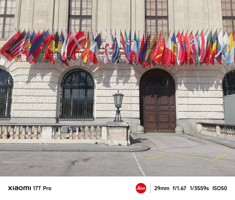 A large building with many international flags displayed above arched windows and a tall wooden door, photographed on a sunny day. Camera specs and branding appear at the bottom of the image.