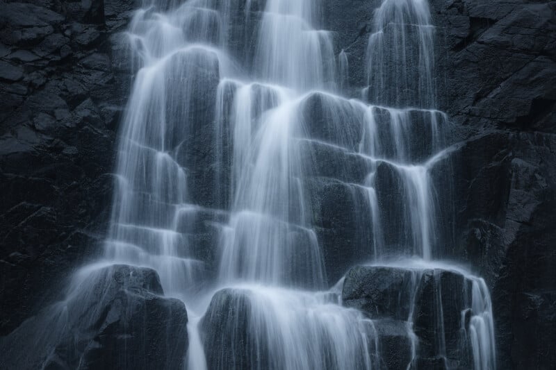 A close-up view of a cascading waterfall flowing over dark, rugged rocks, captured with a long exposure to create a smooth, misty effect on the water.