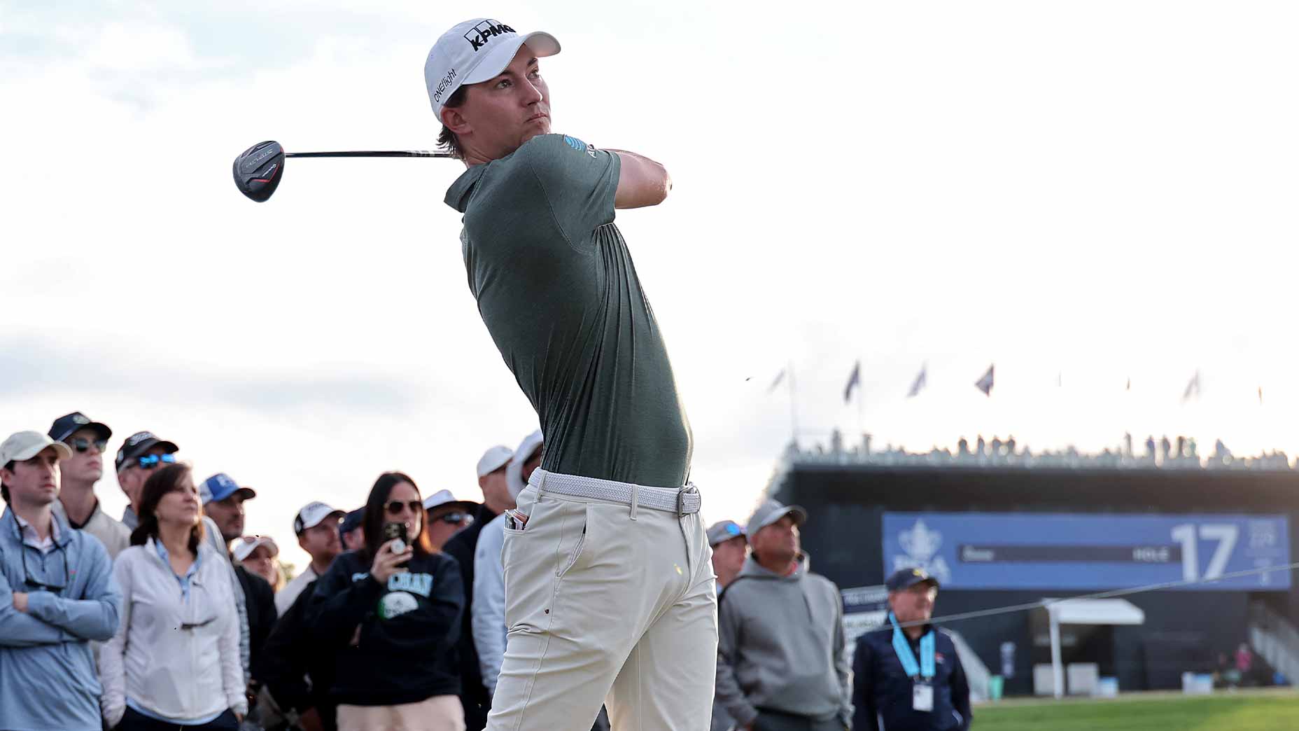 A golfer in a gray shirt and white pants swings a golf club at the PGA Championship, watched by a crowd of spectators. In the background, a large blue scoreboard and a stand filled with people can be seen.