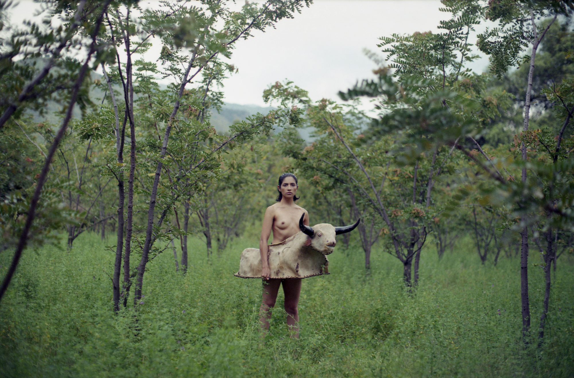 A topless woman stands in a lush green orchard, holding a large sculpted bull figure at waist height and staring directly at the camera.