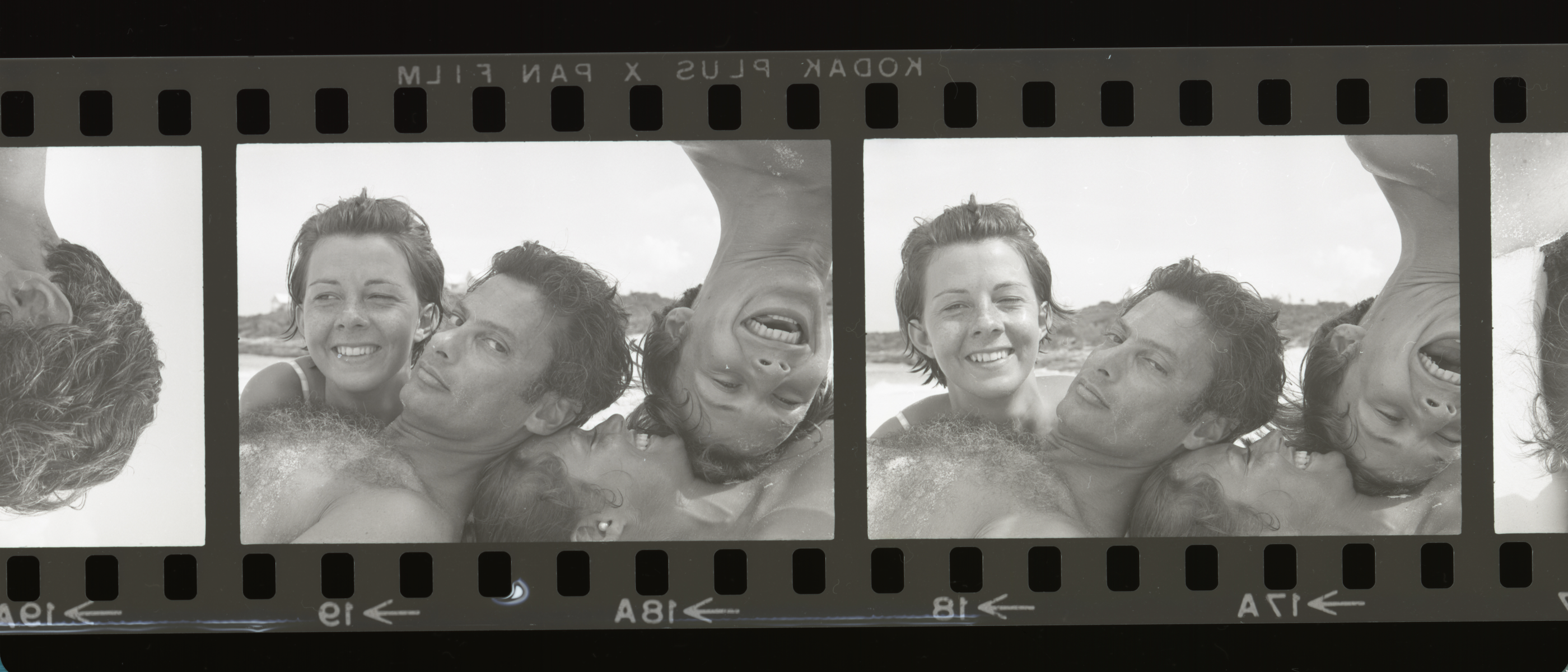 A strip of Kodak film frames showing Avedon and Lauren Hutton pressed cheek to cheek with a third person on a sunlit beach, laughing and tumbling into the camera.