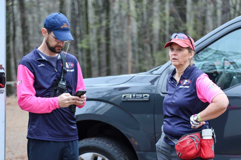 A man and a woman in matching navy blue shirts with pink sleeves stand ouside an event, gear on hand
