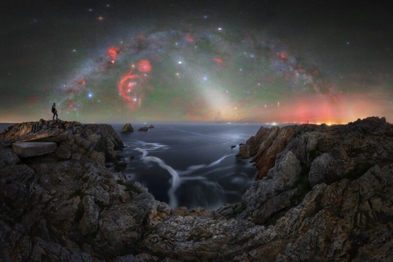 A man stands at the top of a cliff overlooking the sea and the Milky Way arches overhead.