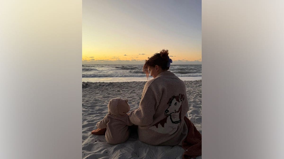Millie Bobby Brown sits on the beach at sunset beside her daughter while looking out toward the ocean.