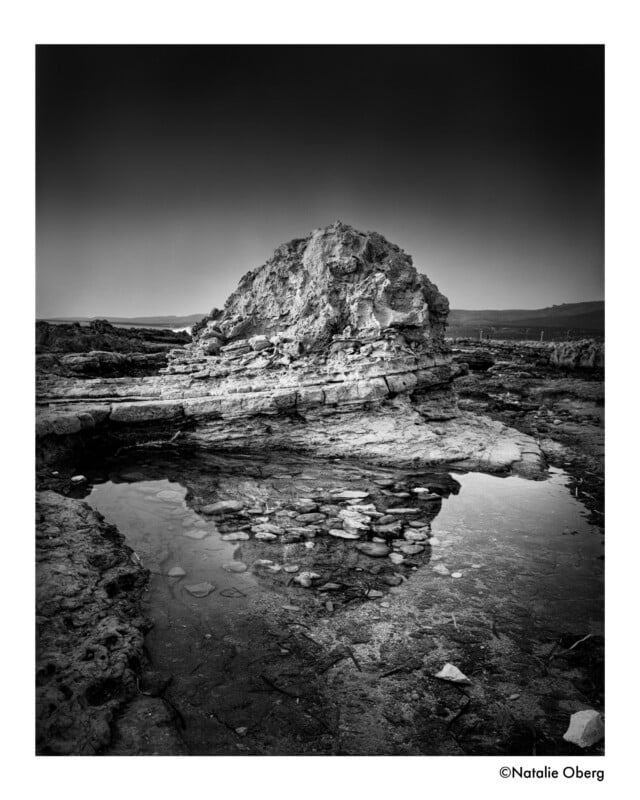 Black and white photo of a large rocky formation reflected in a shallow tidal pool, with rugged terrain and a distant horizon under a dark, dramatic sky.