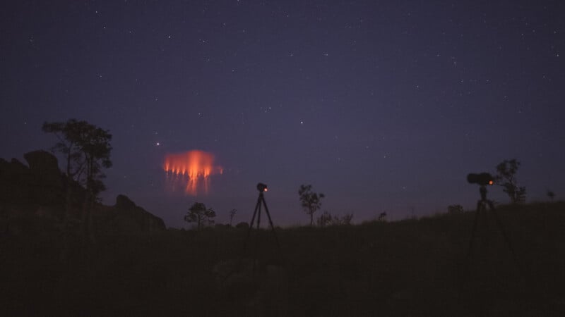 A dark, starry night sky with a distant red lightning phenomenon called a sprite glowing above the horizon. Silhouettes of trees and two tripods with cameras are visible in the foreground.