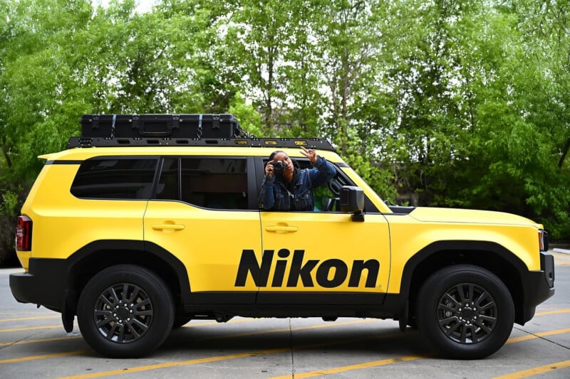 A person is taking a photo from inside a bright yellow SUV with the word "Nikon" in large black letters on the side. The vehicle is parked outdoors, surrounded by green trees.