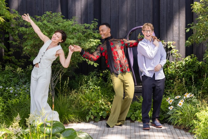 Emma Willis, Josh Widdicombe and Johannes Radebe pose in a garden. Emma holds hands with Johannes, who has his hand over Josh's shoulder, as they pose