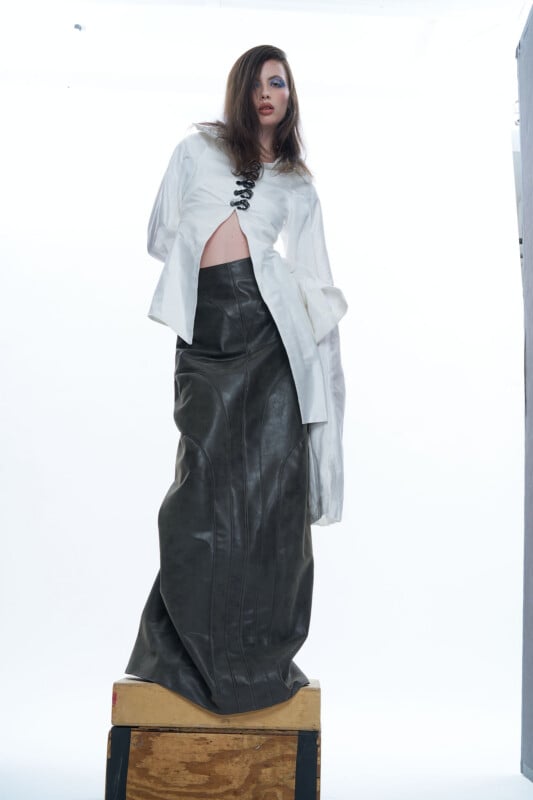 A person stands on a wooden box against a white background, wearing a loose white shirt partially open at the front and a long, high-waisted, dark leather skirt with vertical seams. Their hair is styled to one side.
