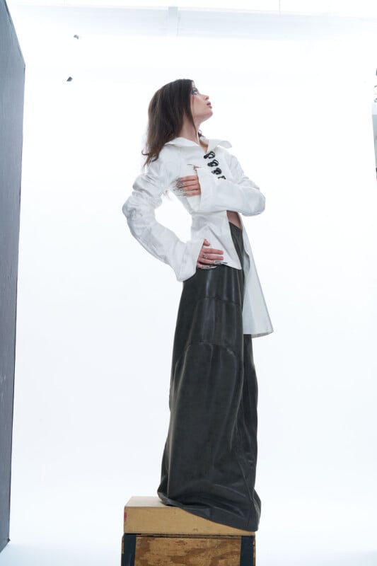 A woman stands on a wooden box, wearing a white button-up shirt and wide-leg black pants. She looks up to the side with her hands on her hips, against a bright white background.