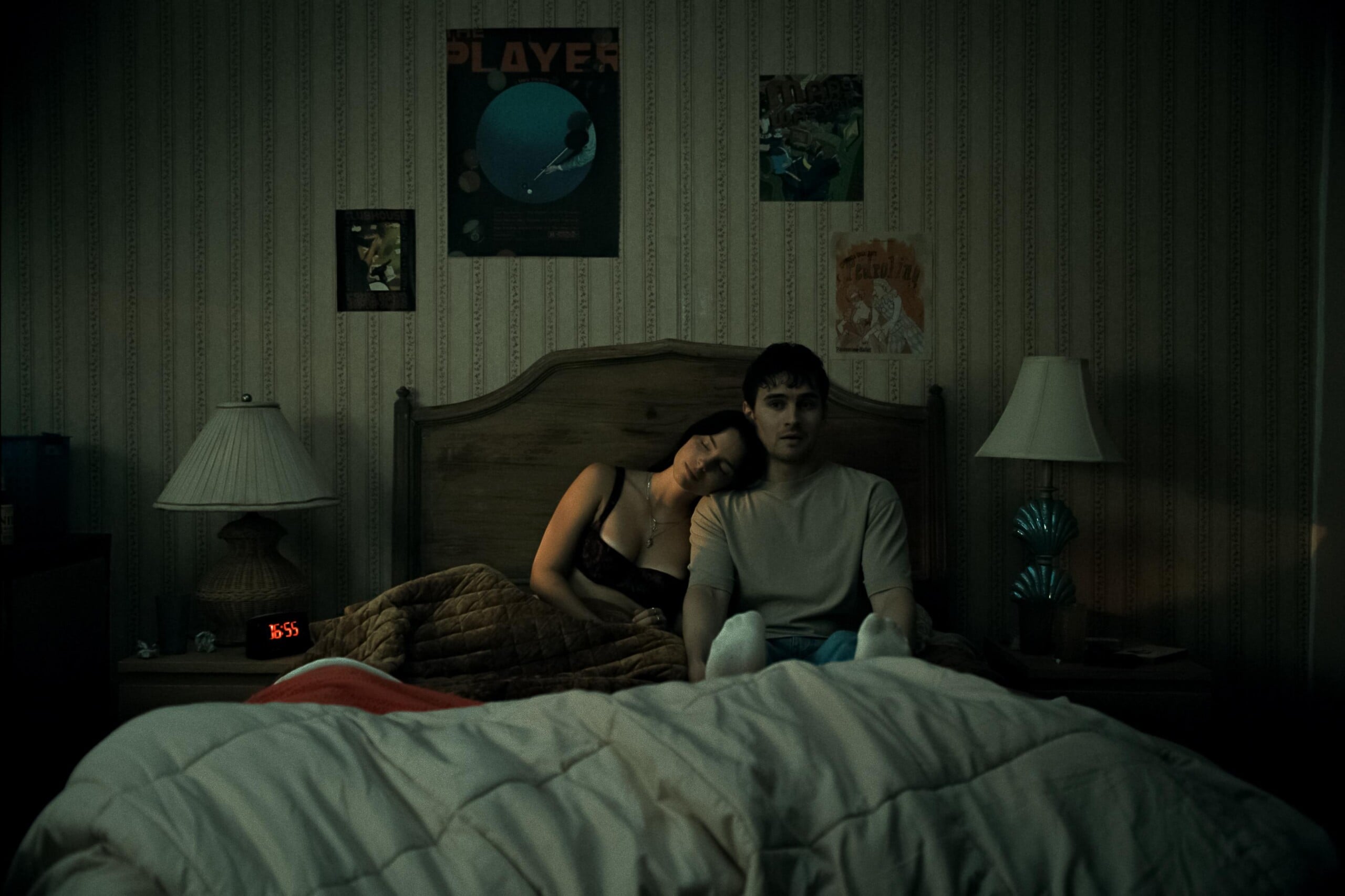 A woman rests her head on a man's shoulder as they sit on a bed in a dimly lit bedroom, surrounded by posters and lamps on bedside tables. The atmosphere feels quiet and contemplative.