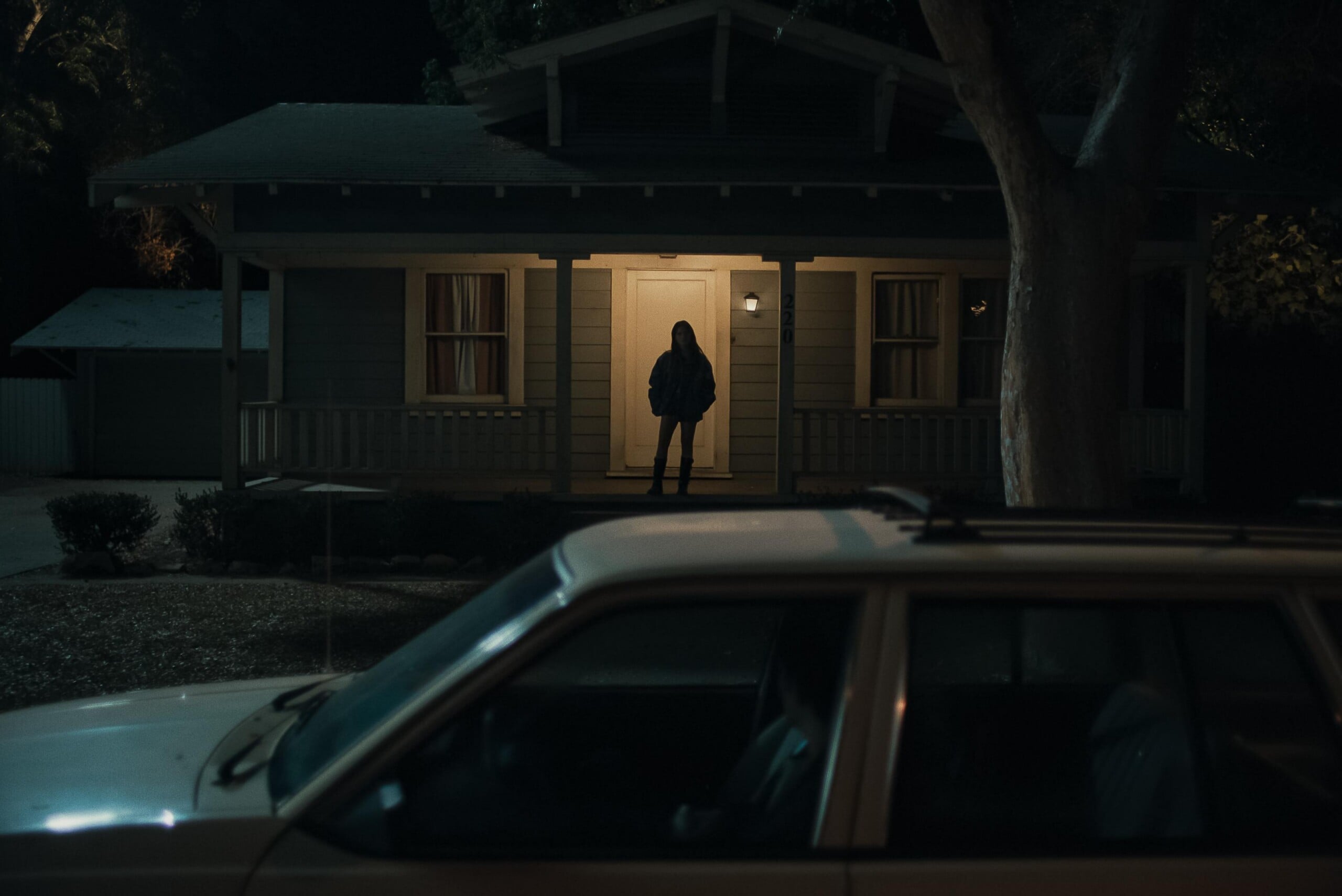 A person stands silhouetted in the light of a front porch at night, facing the door of a house, with a white car parked in the foreground and trees partially obscuring the scene.