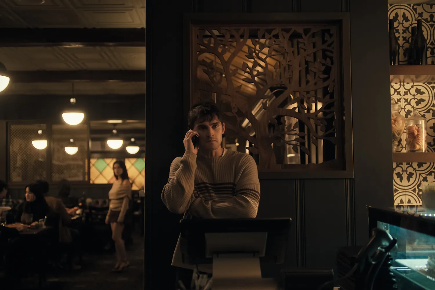 A man in a beige sweater stands at a restaurant counter, talking on the phone. Warm lighting and decorative wall panels create a cozy atmosphere. Diners are seated in the background, and a woman in a beige outfit stands nearby.