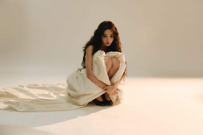A woman with long dark hair sits on the floor, wrapped in a large white cloth. She is looking forward with a serious expression, surrounded by soft, neutral lighting in a minimal studio setting.