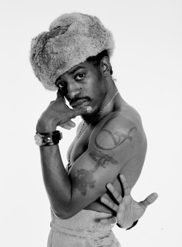 A shirtless man poses with his arm flexed, wearing a large fur hat and jewelry. He looks directly at the camera, touching his face with one hand. His arm shows visible tattoos. The background is plain white.