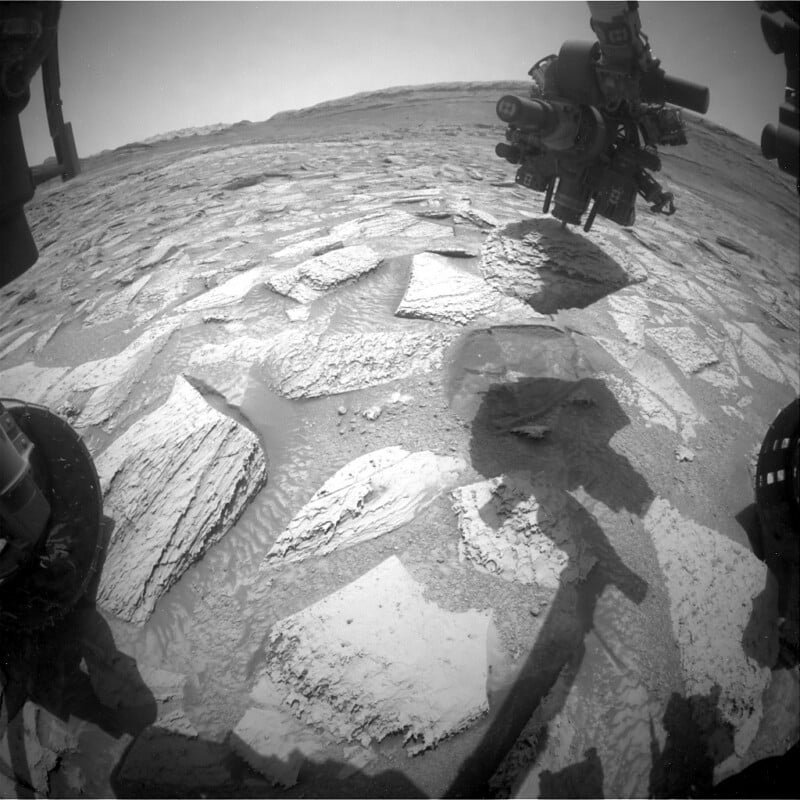 Black-and-white image showing rocky terrain with flat, layered stones on Mars. A robotic arm and wheel from a rover are visible, along with the rover's shadow cast on the ground. The horizon is faint in the distance.