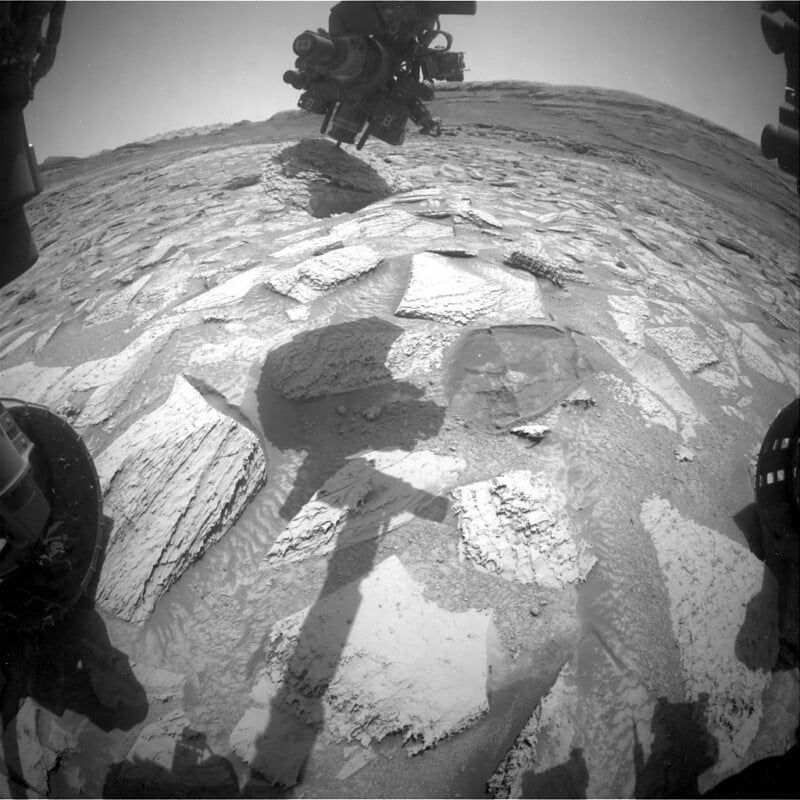 Black and white image from a Mars rover shows rocky, uneven terrain with layered flat stones. Parts of the rover, including its arm and wheel, are visible, casting shadows on the Martian surface under a clear sky.