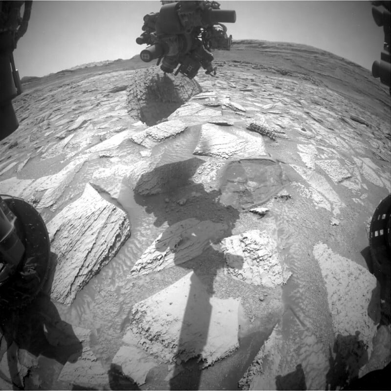 Black and white image from a Mars rover shows rocky, cracked terrain with the rover's shadow and part of its mechanical arm visible, as well as distant hills on the horizon under a clear sky.