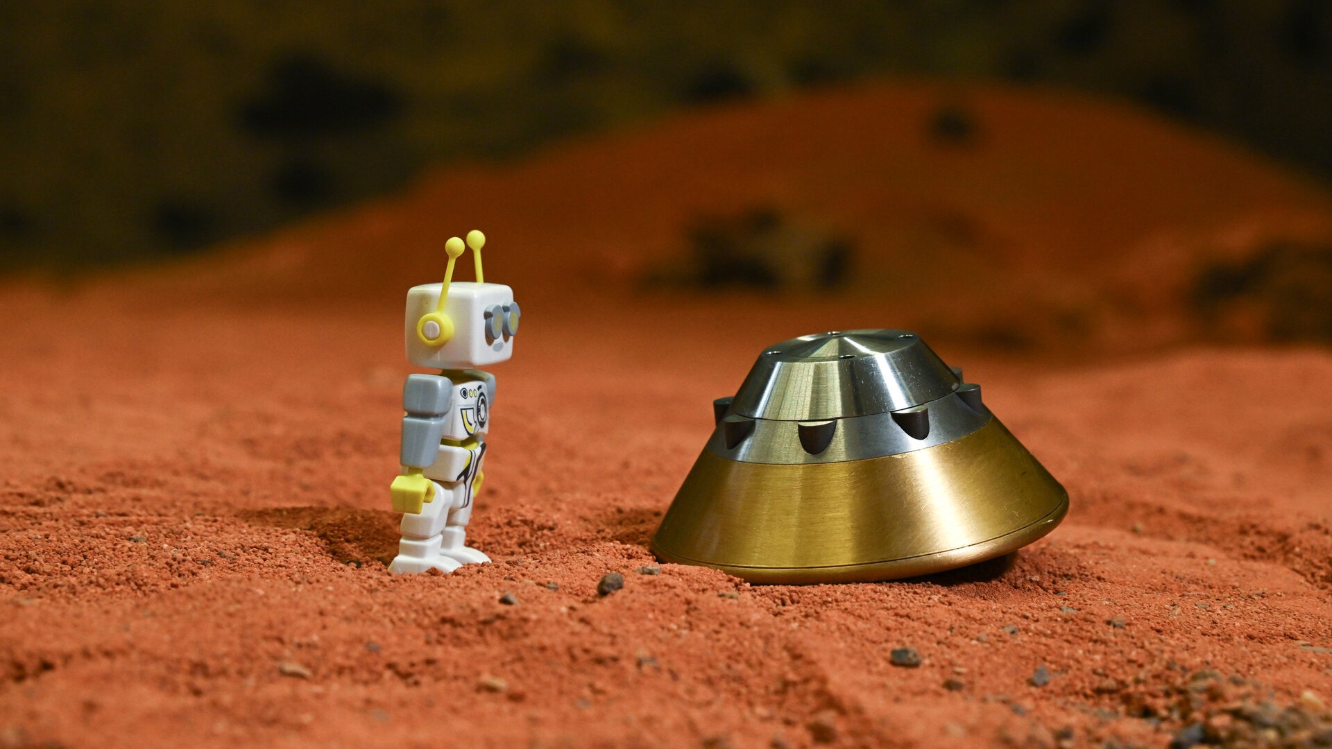 A small robot Lego figurine stands to the left of a miniature space capsule, both sitting in reddish dirt.
