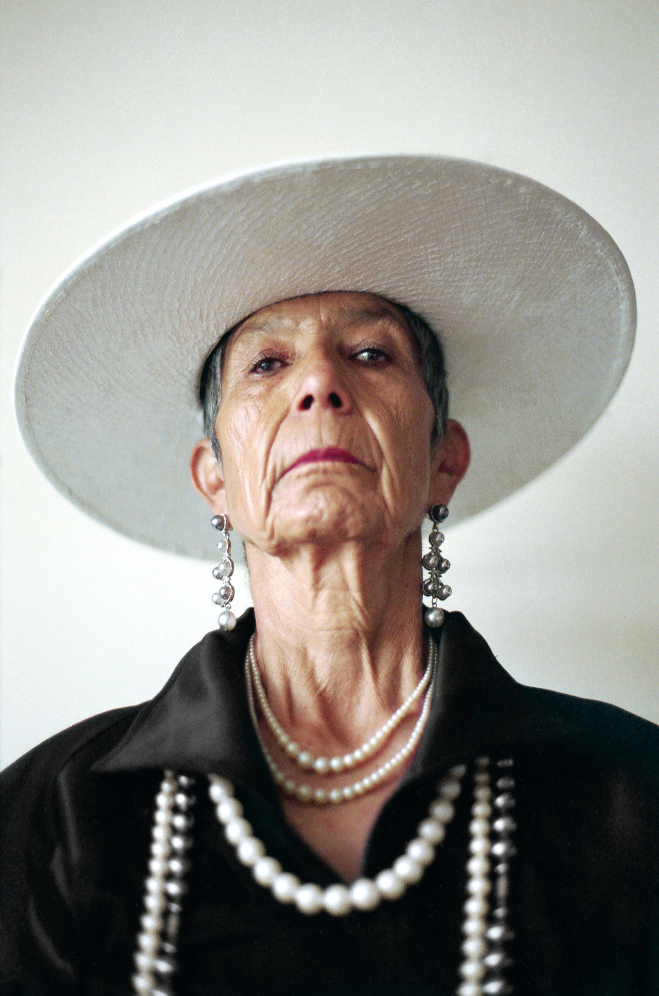 A close-up portrait of an older woman wearing a wide-brimmed white hat, multiple strands of pearls and long drop earrings, photographed in black against a plain off-white background.
