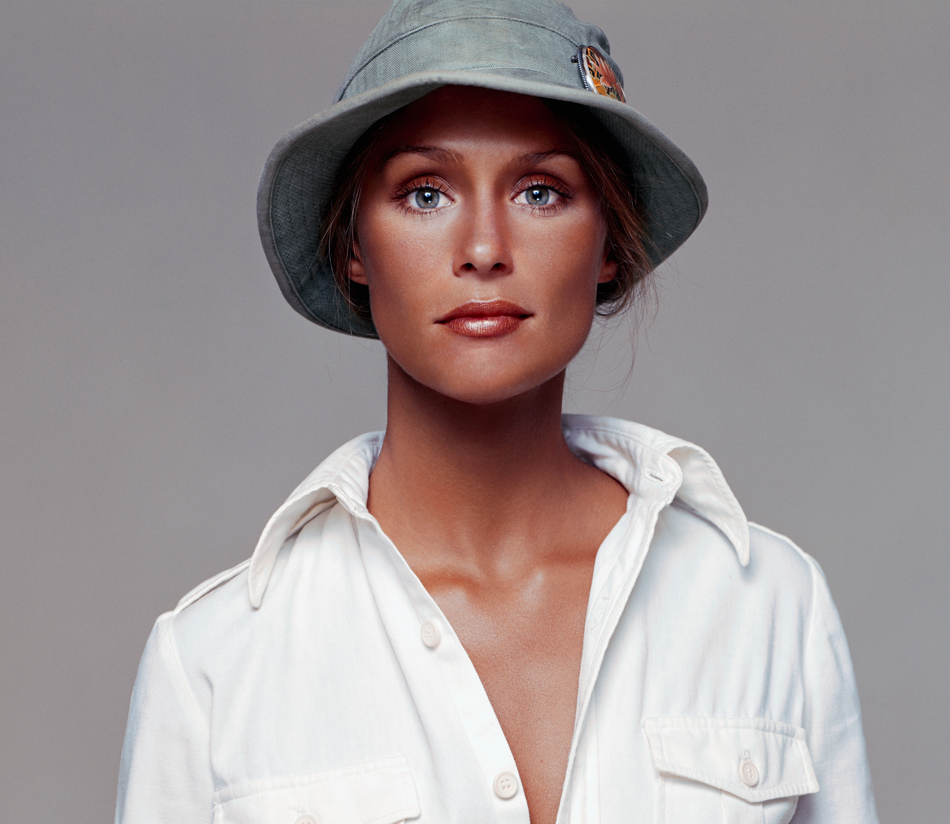 A colour portrait of Lauren Hutton in a soft grey bucket hat and open white shirt, her blue eyes meeting the camera with calm directness.
