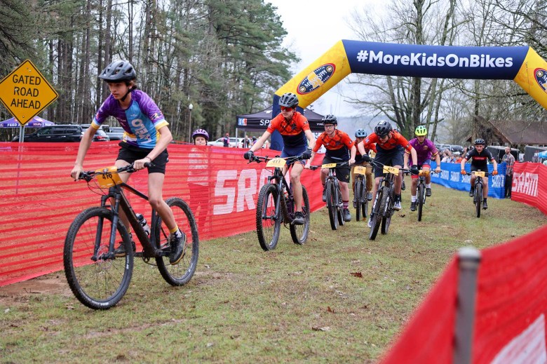 Kids ride on bikes during an outside race event. A over-the-track sign reads #MoreKidsOnBikes