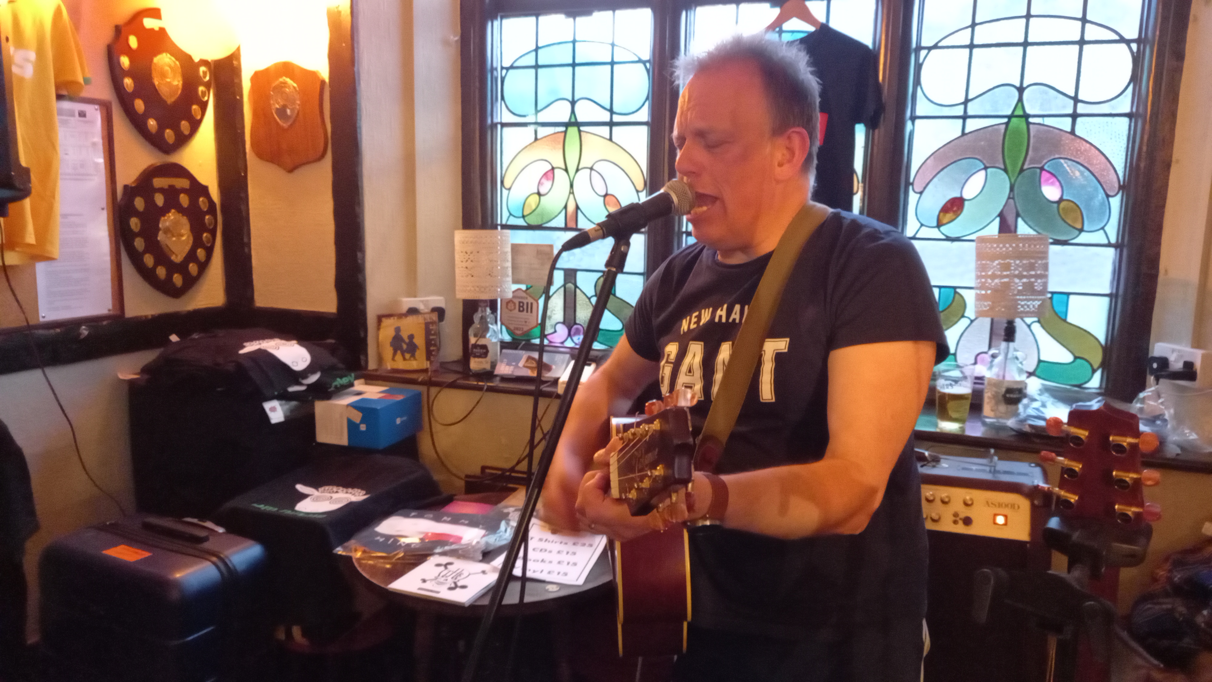 A man in a dark t-shirt sings into a microphone while playing an acoustic guitar in a pub setting with stained-glass windows in the background.