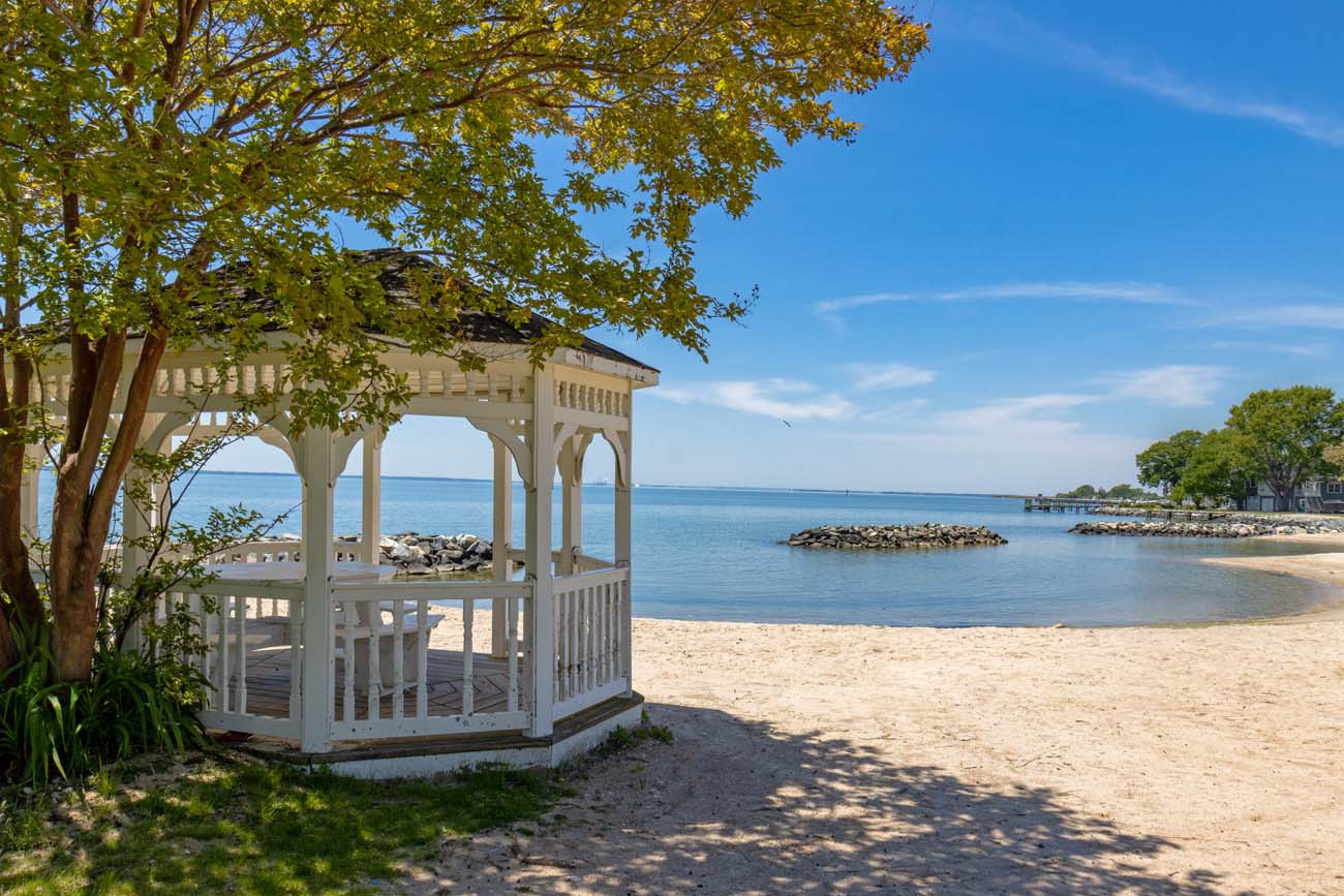 The white octagonal wooden gazebo at Ferry Park Beach in Rock Hall, Maryland, situated at the edge of a sandy bay beach with calm flat blue Chesapeake Bay water stretching to the horizon, a rocky jetty extending into the water, a wooden pier visible to the right, and a tree with fresh spring foliage shading the gazebo from a clear blue sky
