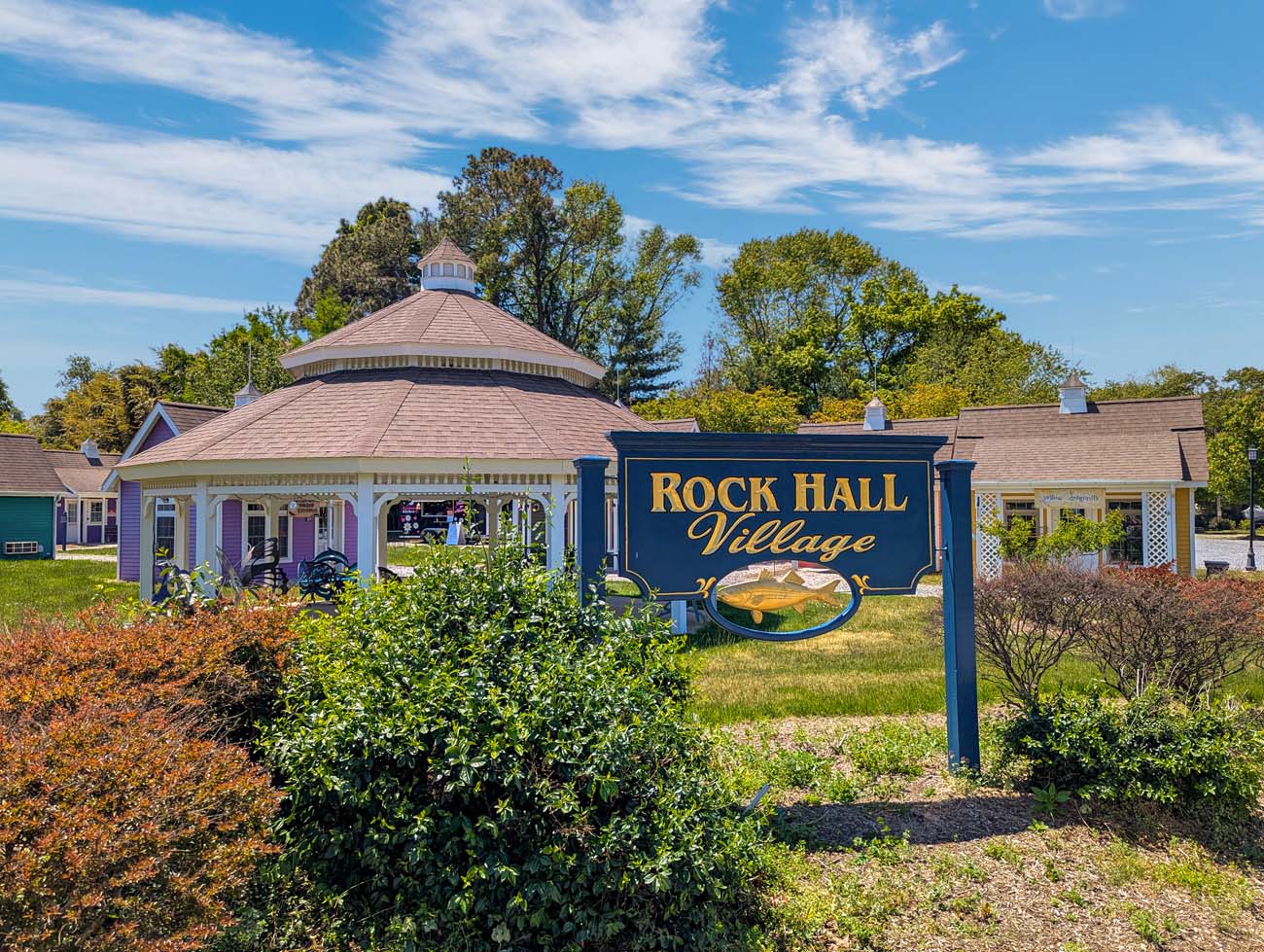 The Rock Hall Village entrance in Rock Hall, Maryland, featuring a navy blue and gold painted wooden sign reading Rock Hall Village with a decorative rockfish illustration, set in front of a large octagonal gazebo with a conical hip roof, a purple cottage with white trim, yellow and green cottages visible to the right, and mature trees under a bright blue spring sky with wispy clouds