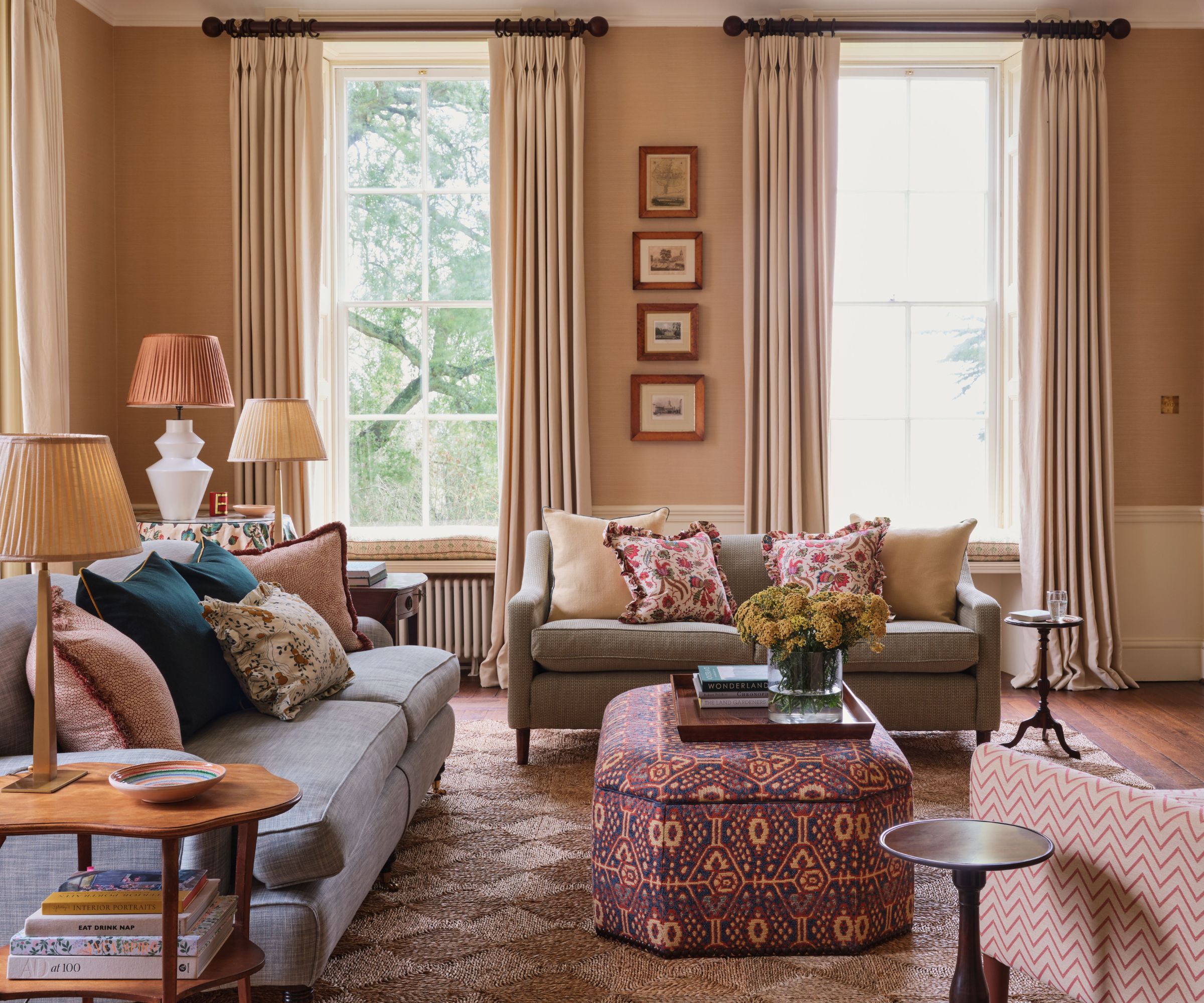 Living room with beige walls, tall windows with heavy cream drapes, a green sofa with floral cushions, a pale blue sofa with colourful cushions, an upholstered ottoman, and a jute checkerboard rug