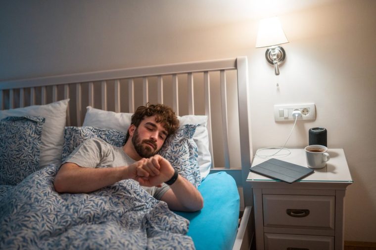 Caucasian male lying in bed, looking at wristwatch in a dimly lit bedroom, evoking sleep issues. Scene includes casual night attire and a bedside table with a cup and book, highlighting restlessness.