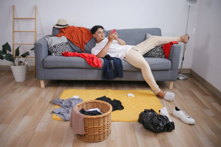 Young Man Relaxed On Sofa While Looking At Smartphone On Messy Room