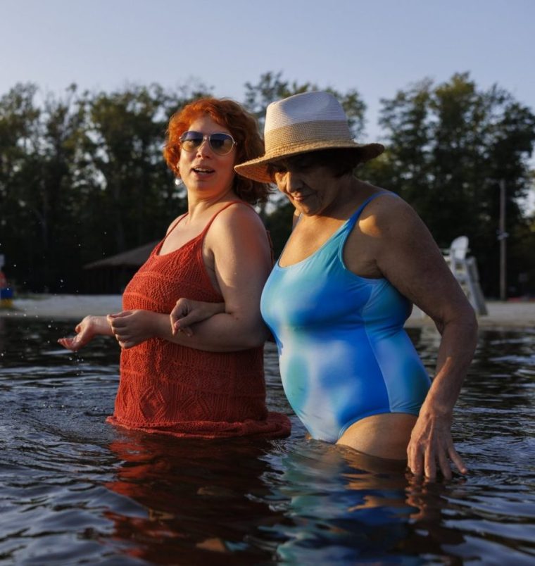 Mid adult redhead woman and her senior mother standing arm in arm waist-deep in lake water. The daughter splashes water with her hand while looking at the camera. The mother wears a sun hat and looks down at the water. They are enjoying a summer afternoon at a sandy beach in the Poconos, Pennsylvania. Trees line the distant shore under a clear sky.