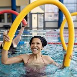 Healthy senior woman doing swim noodle exercises in aerobics pool class. Active senior people doing workout in swimming pool holding swim noodles.