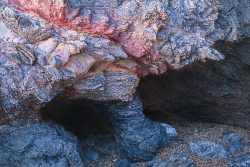 Rock formation with textured, layered surfaces in shades of blue and gray, streaked with reddish and pinkish veins. A small dark cave-like opening is visible at the base, surrounded by loose rocks and gravel.