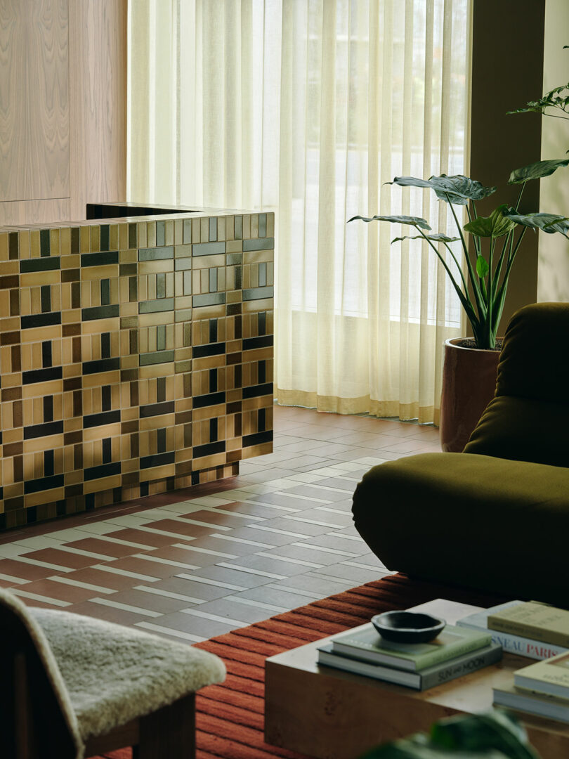A modern living room with a geometric tiled partition, large potted plant, green armchair, coffee table with books, and floor-to-ceiling sheer curtains letting in natural light.