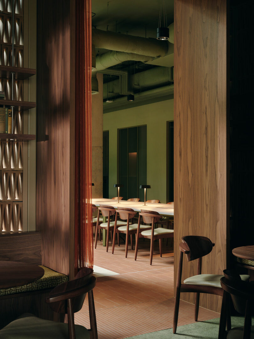 A view into a modern dining area with wooden tables and chairs, soft lighting, and exposed ceiling pipes.