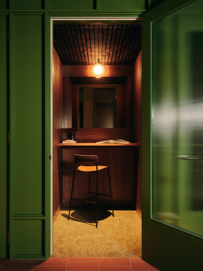 A small room with wood-paneled walls, a high table with an open book, a single black bar stool, warm lighting, and a green door partially open.