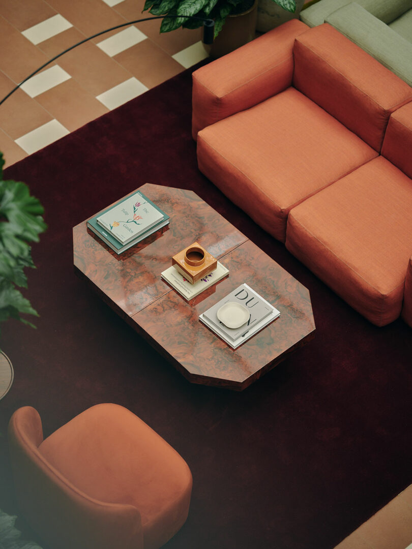A geometric coffee table with books and a ceramic cup sits on a dark rug, surrounded by orange sofas and a peach armchair in a tiled room with potted plants.