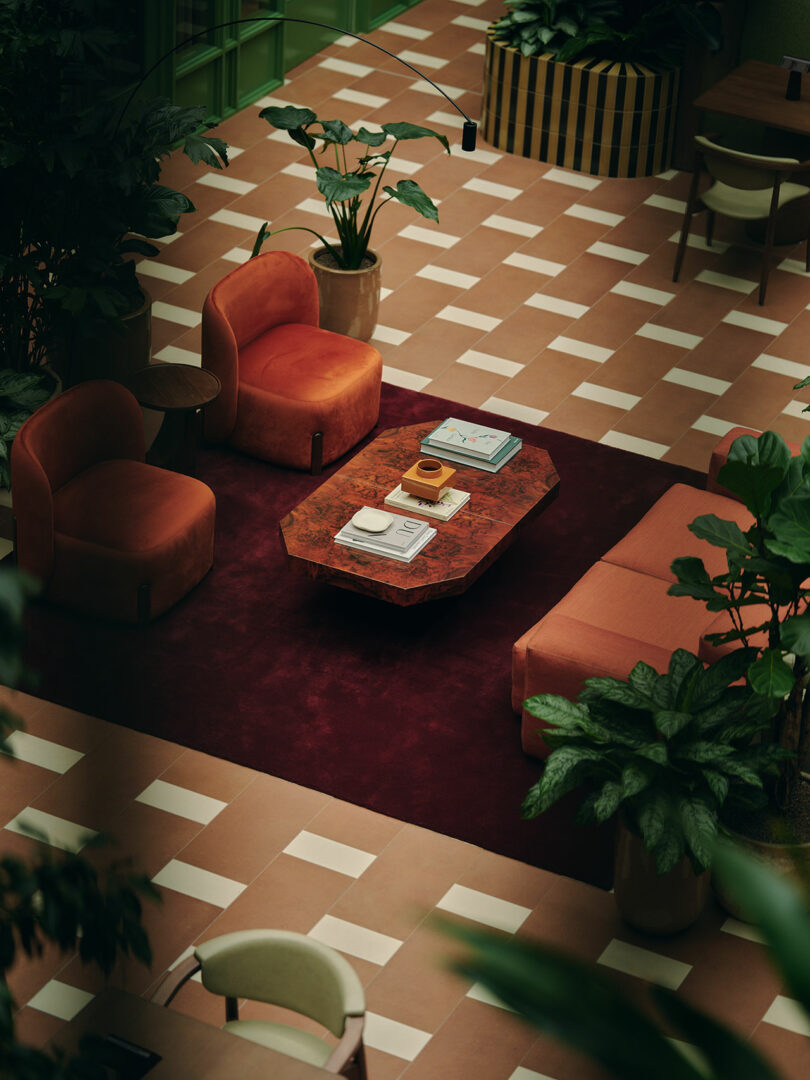 A modern lounge area with orange chairs, a matching sofa, a maroon rug, a wooden coffee table with books, surrounded by potted plants on a checkered tile floor.