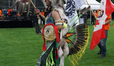 An Indigenous dancer in full regalia — wearing a wolf headdress, colourful ribbon skirt with green and yellow fringe, eagle feather bustle, and hand-painted bag — stands on a grass field holding a ceremonial staff with feathers. A white First Nations flag and a Canadian flag are visible behind them, with a stage and orange-shirted participants in the background. The ISS of BC 'Welcoming Newcomers' logo appears in the bottom right corner.