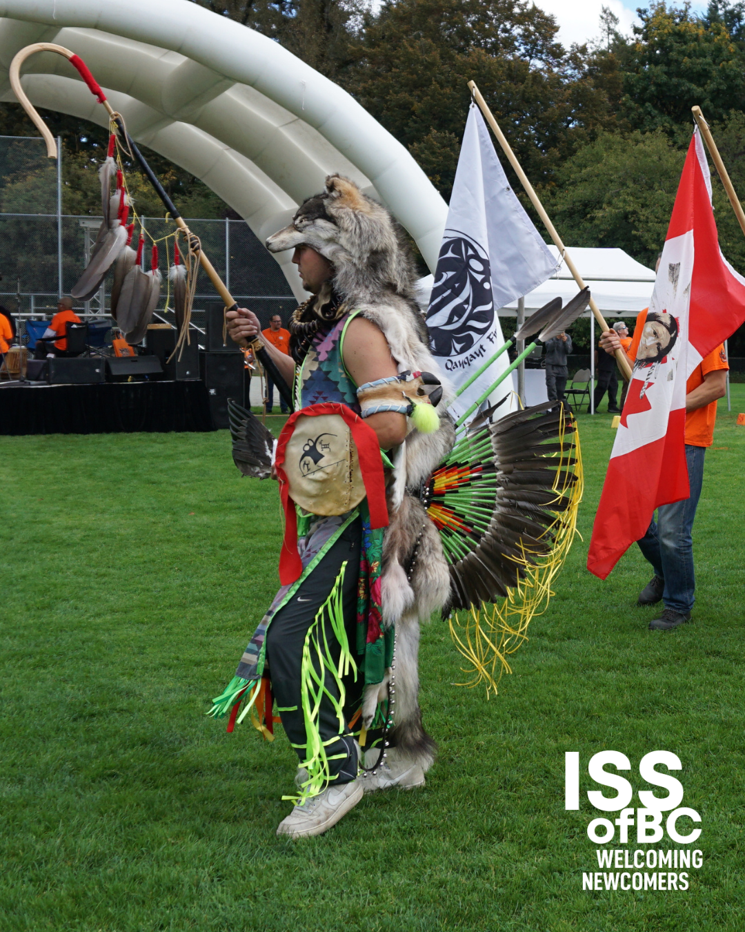 An Indigenous dancer in full regalia — wearing a wolf headdress, colourful ribbon skirt with green and yellow fringe, eagle feather bustle, and hand-painted bag — stands on a grass field holding a ceremonial staff with feathers. A white First Nations flag and a Canadian flag are visible behind them, with a stage and orange-shirted participants in the background. The ISS of BC 'Welcoming Newcomers' logo appears in the bottom right corner.