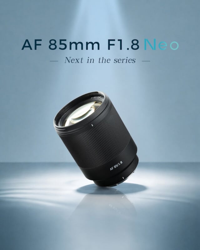A camera lens labeled "AF 85mm F1.8" stands upright on a light blue surface with spotlights. Text above reads "AF 85mm F1.8 Neo" and "Next in the series." The background is soft blue with subtle lighting.