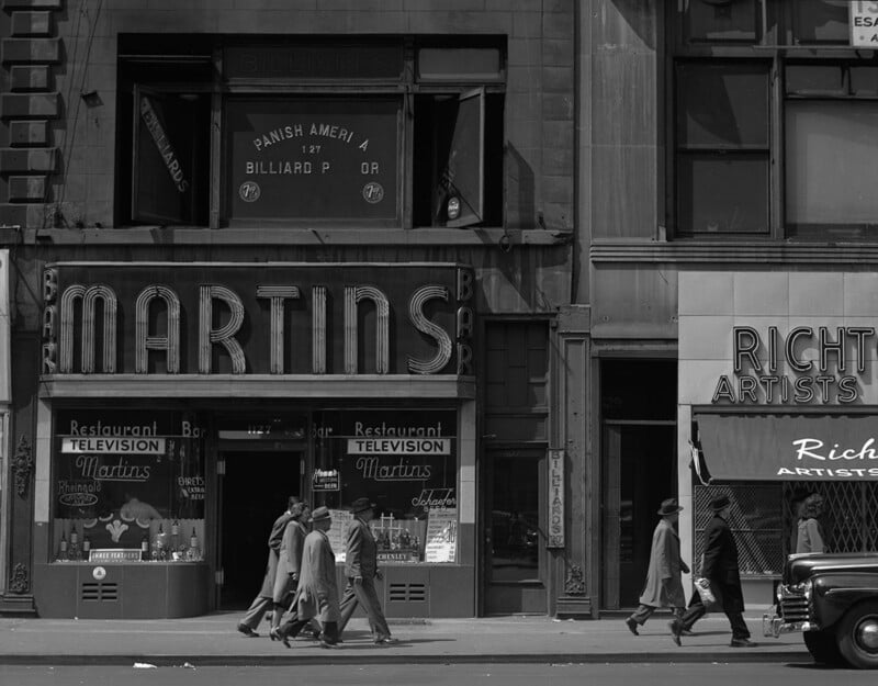 Black-and-white photo of people in coats and hats walking past MARTINS Bar and Restaurant, which advertises television. Above is a sign for a billiard parlor. A vintage car is parked on the street.